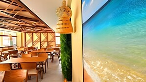 A restaurant with wooden tables and chairs, a hanging light fixture, and a large wall mural of a beach scene. - Makia Beach Experience (Ipojuca)