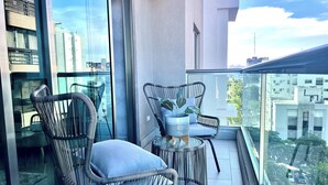 A balcony with wicker chairs and a small table, large windows, and a view of the cityscape.
