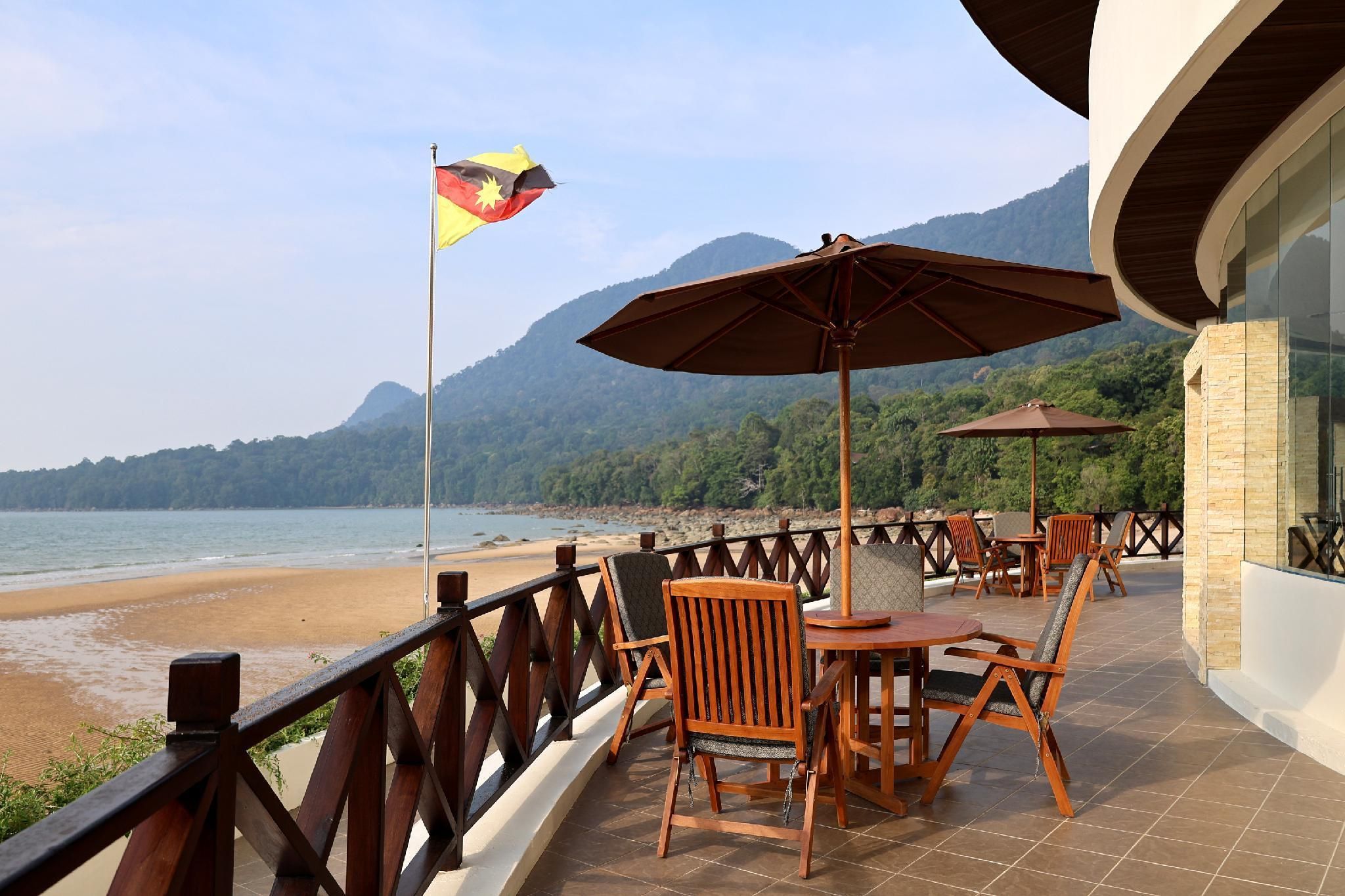 Een terras met houten stoelen en tafels, een vlag en uitzicht op het strand en de bergen.