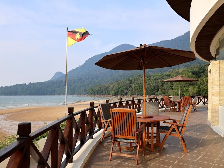 A terrace with wooden chairs and tables, a flag, and a view of the beach and mountains.