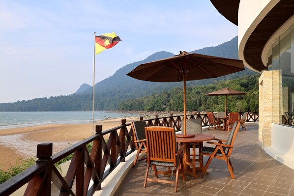 A terrace with wooden chairs and tables, a flag, and a view of the beach and mountains. - Damai Lagoon Resort (Kuching)