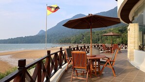 Una terrazza con sedie e tavoli di legno, una bandiera e vista sulla spiaggia e sulle montagne.