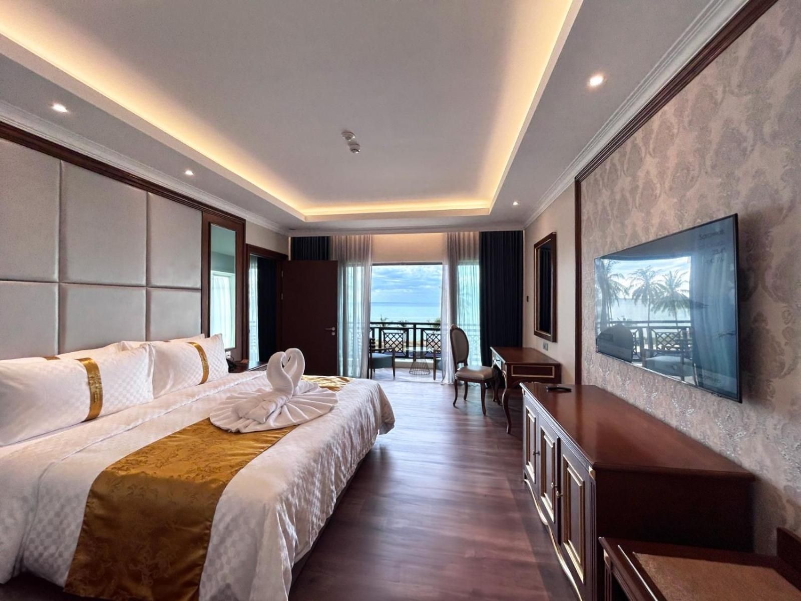 A hotel room with a large bed, a desk, a chair, a TV, and a balcony with a view of the sea.