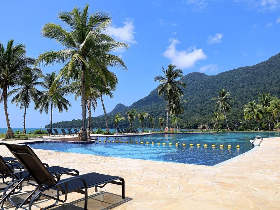 A pool area with lounge chairs and palm trees, surrounded by mountains and a clear sky.