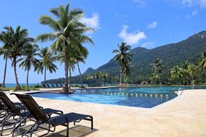 A pool area with lounge chairs and palm trees, surrounded by mountains and a clear sky. - Damai Lagoon Resort (Kuching)