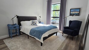 Un dormitori amb llit, una tauleta de nit, una cadira, una finestra amb cortines i un quadre a la paret.