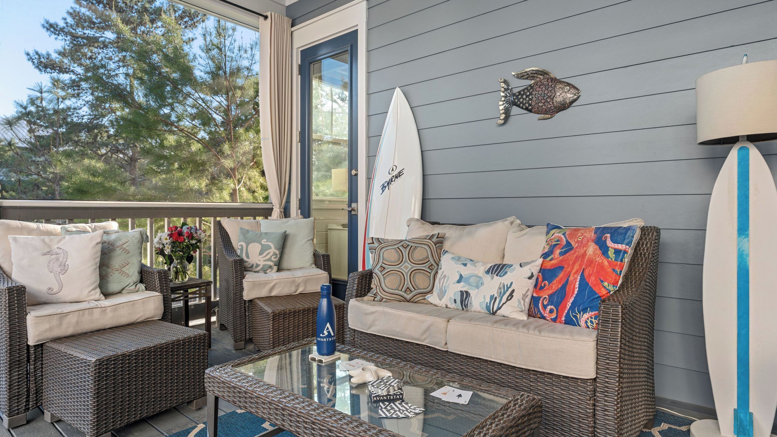 A cozy outdoor seating area with wicker furniture, a glass coffee table, and a surfboard decoration.
