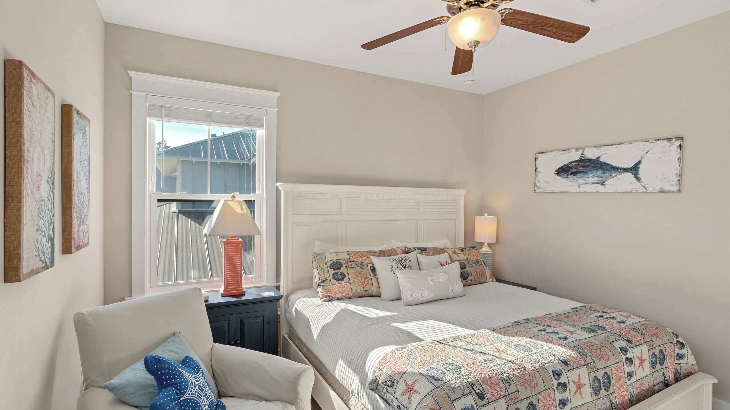 A bedroom with a bed, a chair, a nightstand, a ceiling fan, a window with blinds, and artwork on the walls.