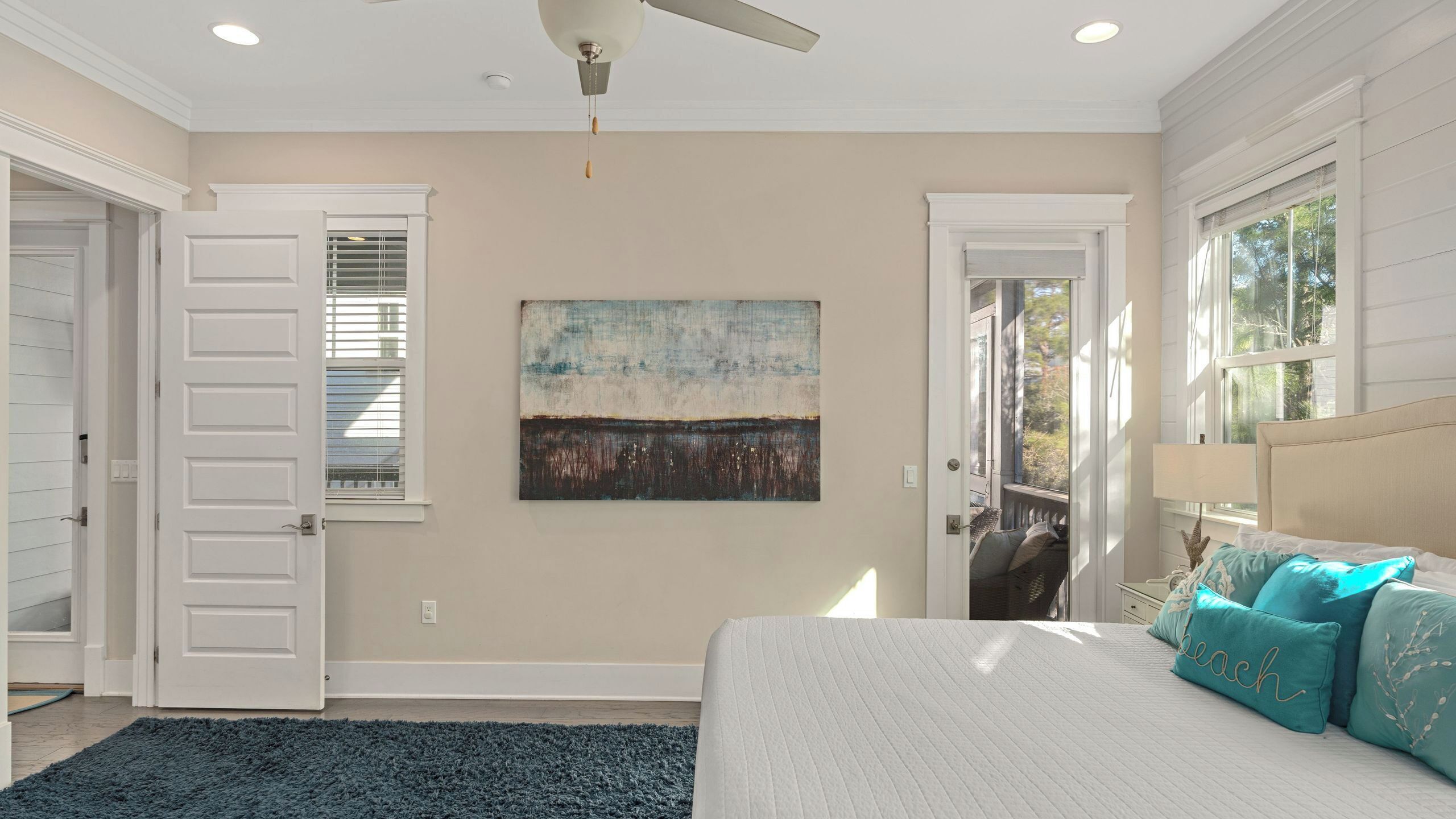 A bedroom with a bed, a ceiling fan, a painting, and a view through a window.