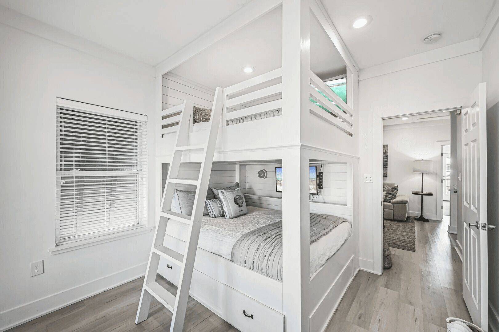 Un lit superposé avec une échelle, une fenêtre avec des stores et une porte donnant sur une autre pièce.