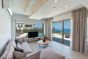 A modern living room with a large TV, a sofa, a round coffee table, and a view of the sea through large windows. - Skyfall Villas - With 12 Bedrooms (Atsipopoulo)