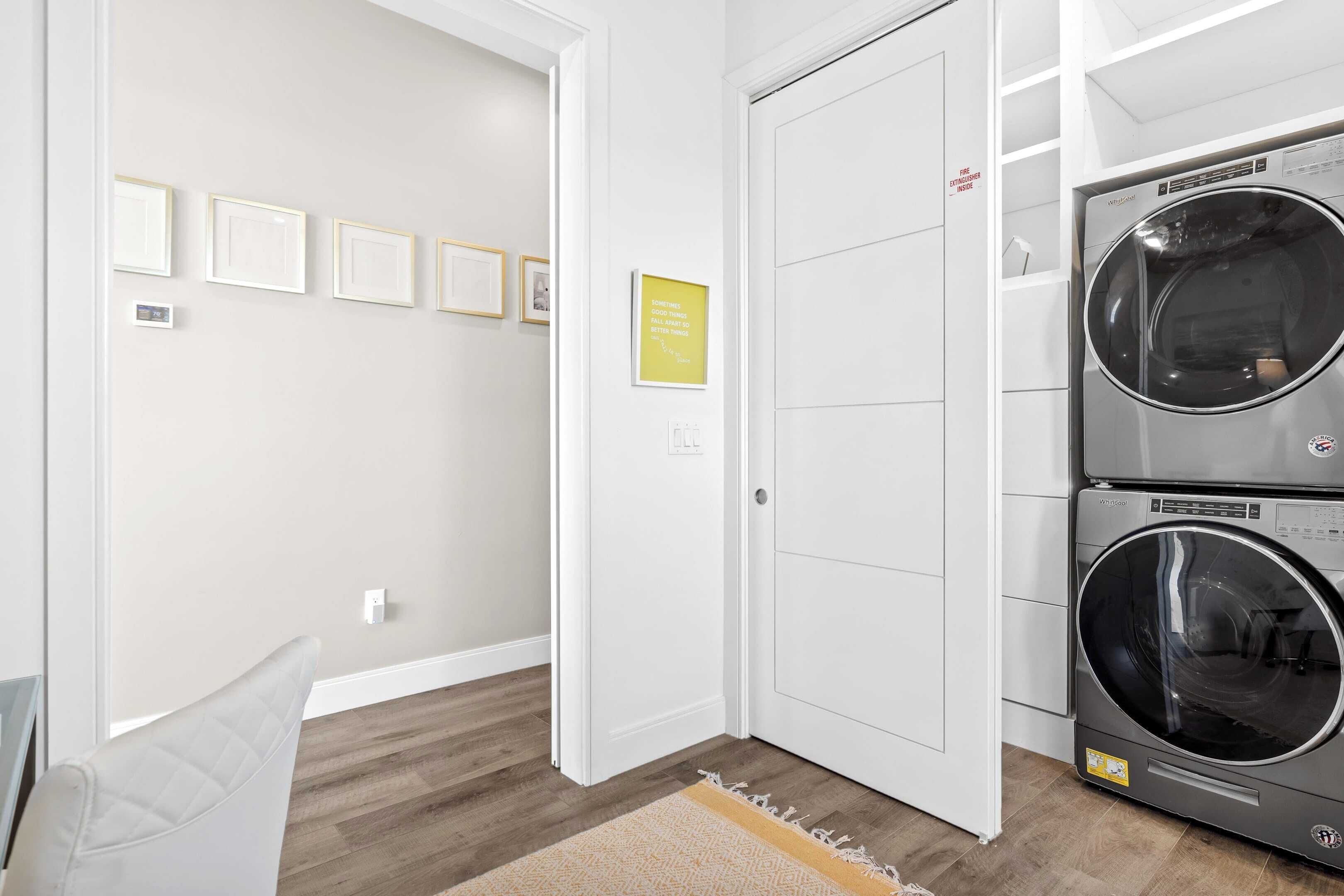 A laundry room with two stacked front-loading washing machines, a door with a sign, and a wall with framed pictures.
