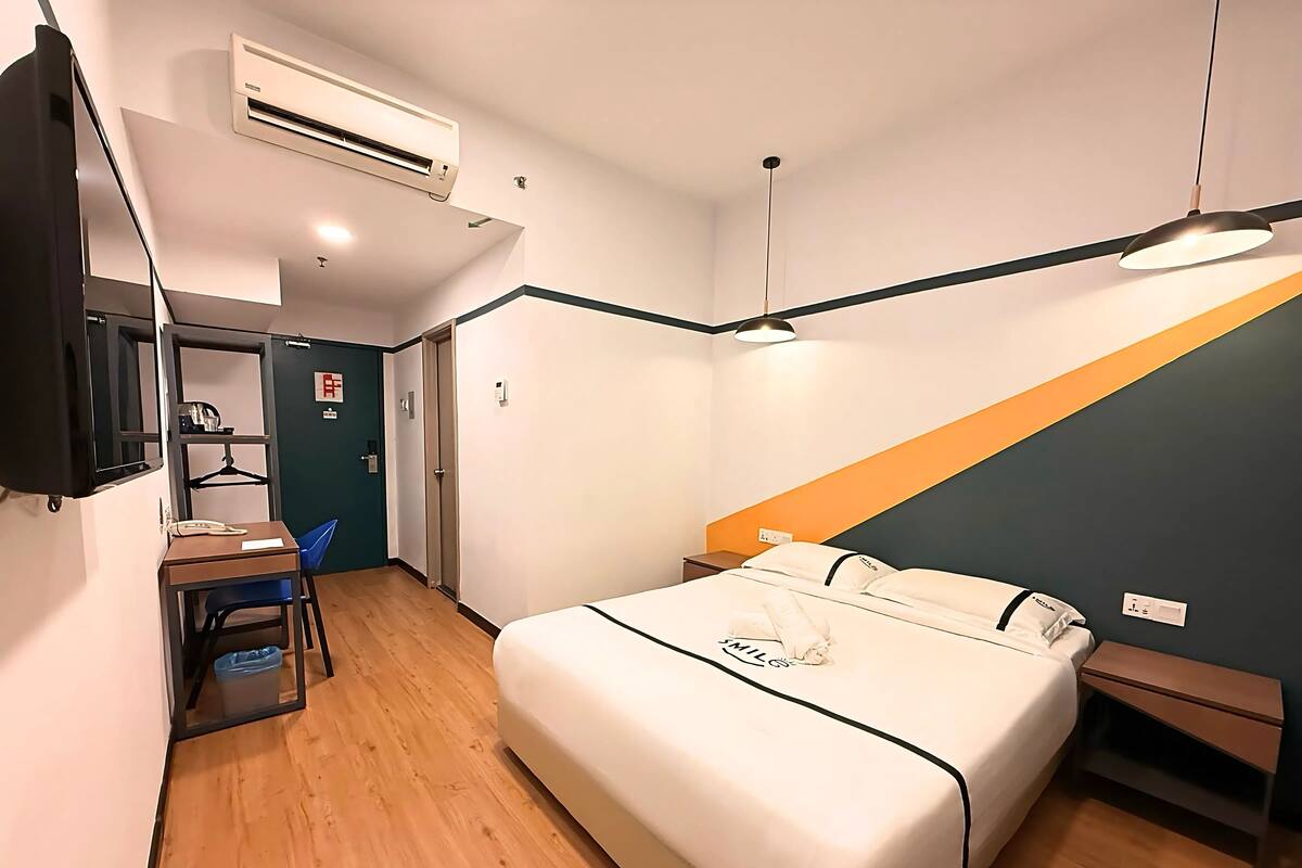 A hotel room with a bed, bedside table, desk with chair, and an air conditioning unit.
