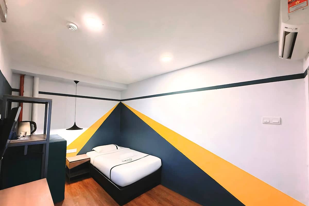 A compact room with a bed, a desk, and a chair. There is a shelf with items on it, and a wall-mounted air conditioner.