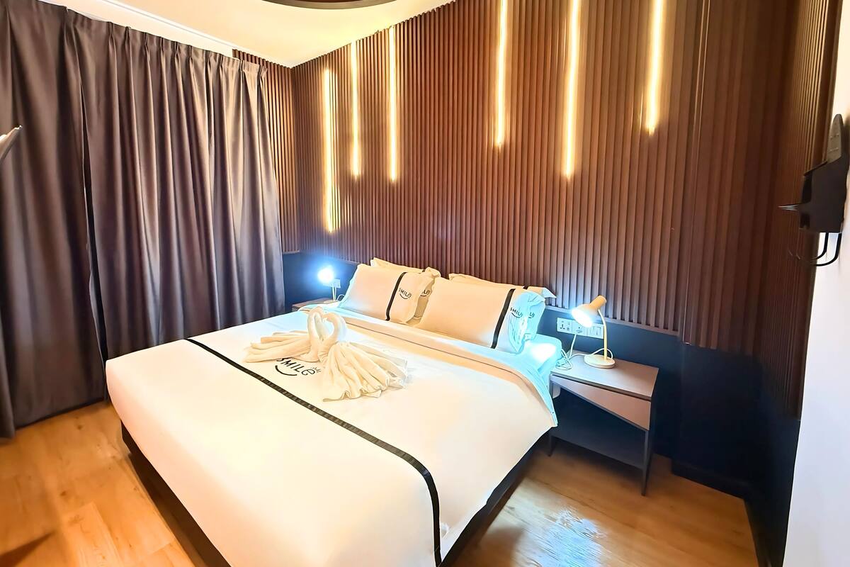 A modern hotel room with a large bed, a bedside table with a lamp, and a wall with vertical blinds.