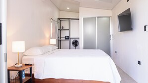 A hotel room with a bed, a bedside table with a lamp, a fan, and a wardrobe.