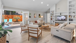 Ein geräumiges Wohnzimmer mit Sofa, Sesseln, Couchtisch und Fernseher. Ein Bücherregal und eine Tür führen auf eine Terrasse mit Holzzaun und Pflanzen.