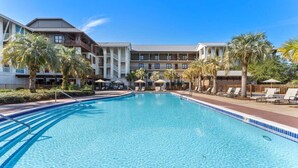 A swimming pool with lounge chairs and palm trees, surrounded by a hotel building with multiple balconies.