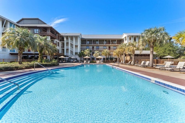 A swimming pool with lounge chairs and palm trees, surrounded by a hotel building with multiple balconies. - Red Fish Blue Mountain by Avantstay Private Beach + Gulf View (Santa Rosa Beach)
