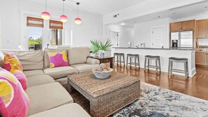 A modern living room with a beige sofa, colorful pillows, a woven coffee table, and a kitchen area with a white countertop and wooden cabinets.