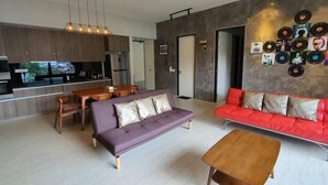 Un saló modern amb un sofà morat, un sofà vermell i una taula de centre de fusta. Hi ha una zona de cuina amb armaris de fusta i una taula de dinar.