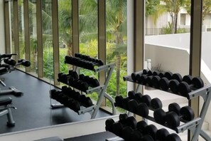 Un gimnasio con mancuernas dispuestas en estanterías, ventanas amplias y vistas al exterior con vegetación.