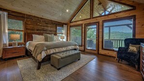 Un dormitorio de cabaña de madera con una cama grande, un banco, una silla y una ventana con persianas.