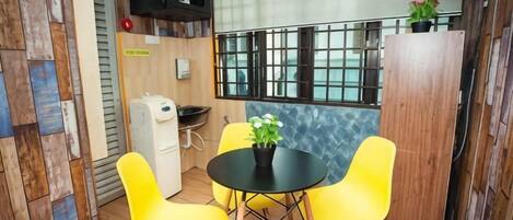 Una habitación pequeña con una mesa y sillas, una ventana con rejas, un refrigerador y una pared de madera con un estante.