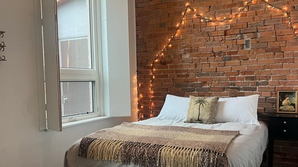 Un dormitorio con pared de ladrillo, cama con ropa blanca, lámpara colgante y ventana con persianas.