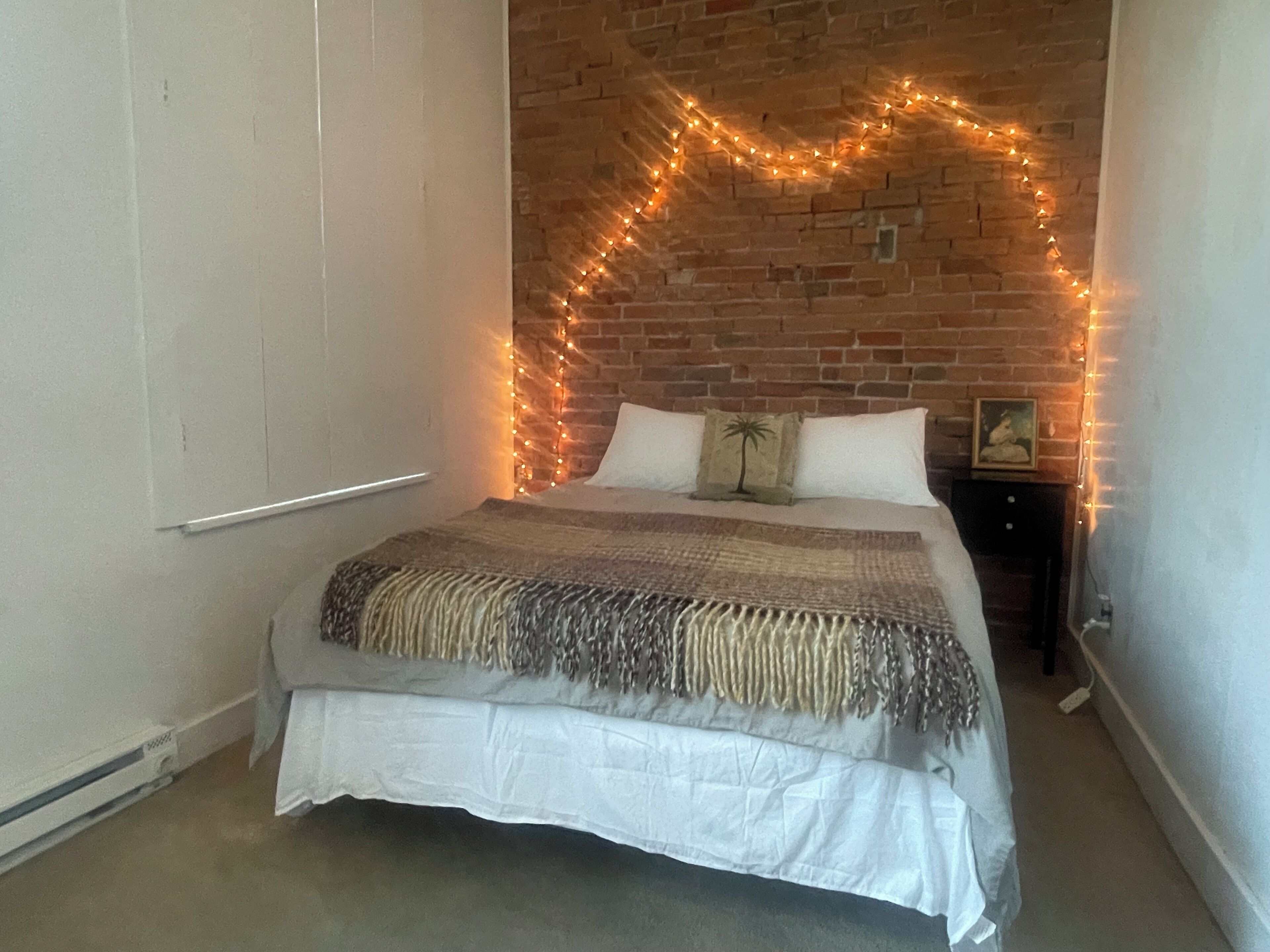 Una camera da letto con parete in mattoni, un letto con coperta bianco e marrone e luci a stringa.