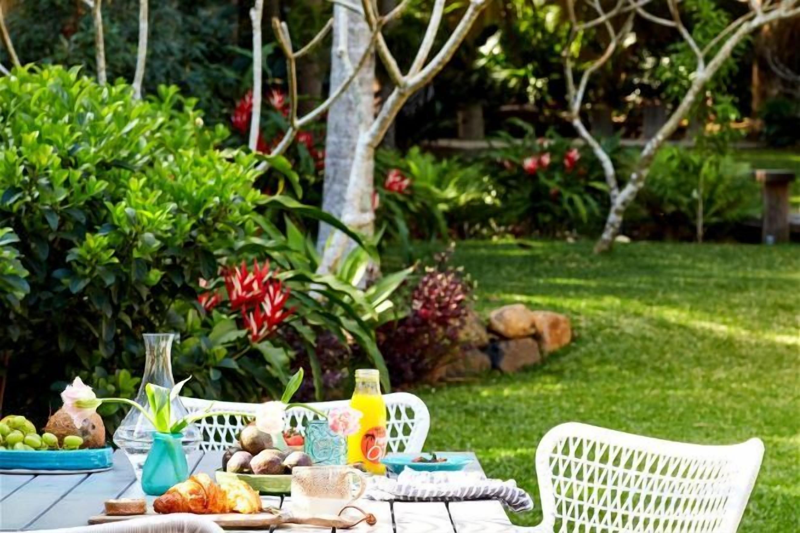 A table set outdoors with a variety of food items and drinks, surrounded by lush greenery and a white chair.