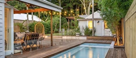 Una piscina con terraza de madera, una zona de estar cubierta y una casa pequeña al fondo.