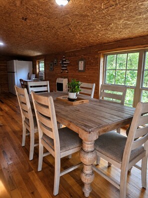 A rustic wooden dining area with a table and chairs, a refrigerator, and a window with a view of greenery.