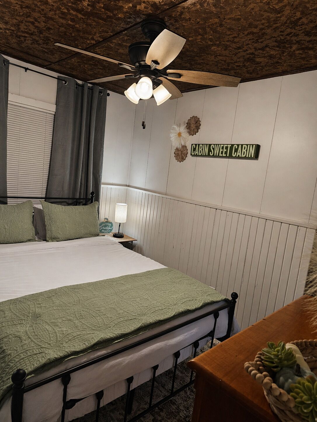 Quarto com cama, ventilador de teto, mesa de cabeceira com abajur, placa com a inscrição 'CABIN SWEET CABIN' e janela com cortinas.