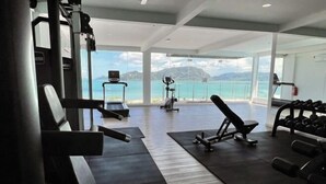 Ein Fitnessstudio mit Cardiogeräten, Krafttrainingsmaschinen und Kurzhanteln, mit Blick auf das Meer und die Berge.