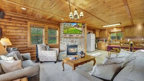 A log cabin interior with a stone fireplace, wooden furniture, and a kitchen area.