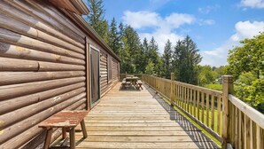A wooden deck with a bench and picnic table, surrounded by a wooden railing, leading to a cabin with a door.