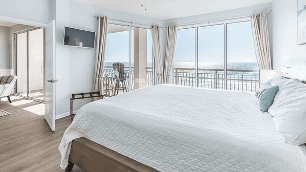 A hotel room with a large bed, a TV mounted on the wall, a balcony with a view of the sea, and a chair by the door.