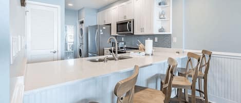 A kitchen with a central island, white cabinetry, a microwave, a refrigerator, and a sink. There are four wooden chairs around the island.