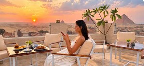 A person seated at a table with food and drinks, overlooking a desert landscape with pyramids in the distance. - Luma Pyramids Boutique Hotel (Giza)