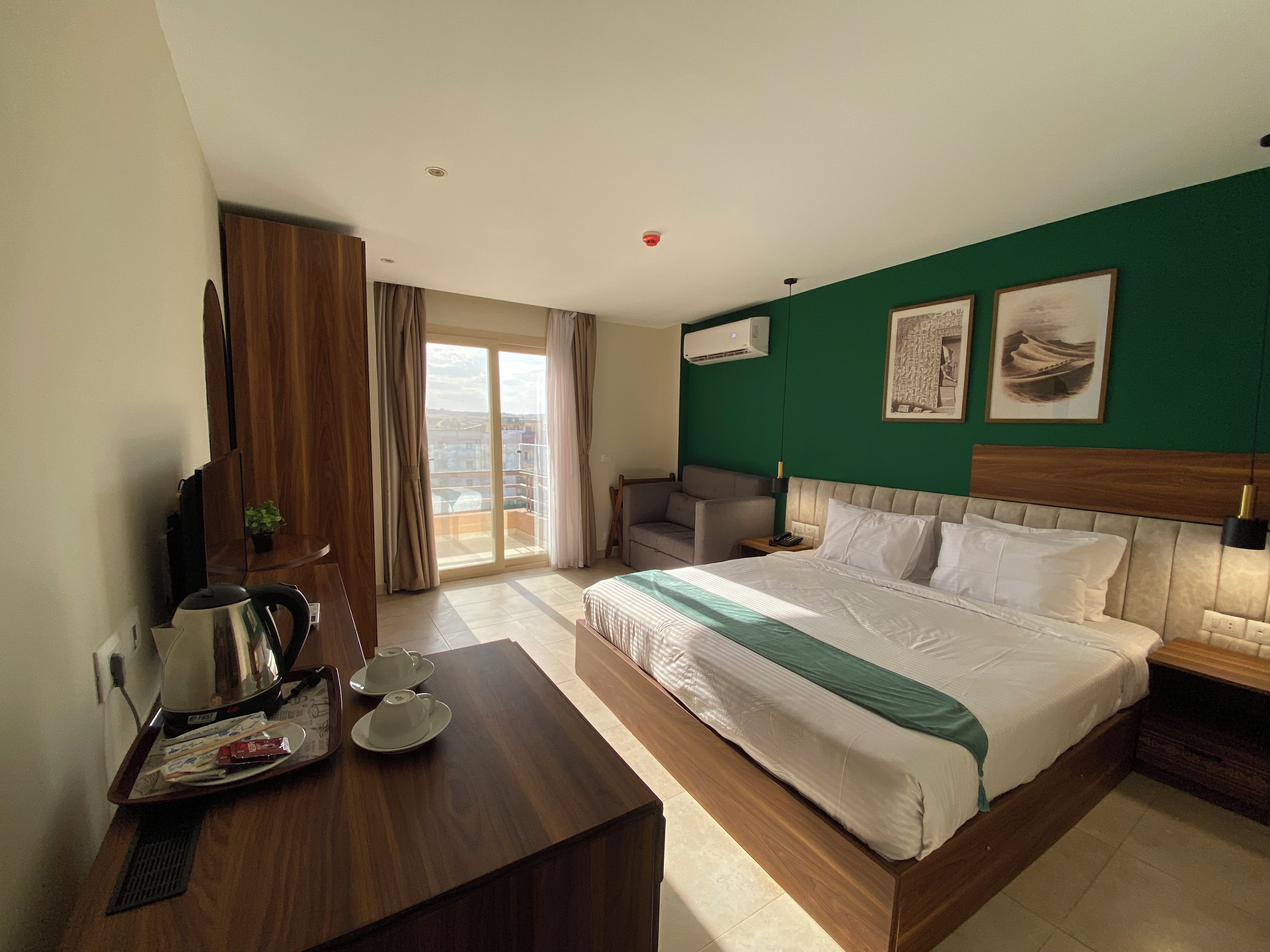 A hotel room with a bed, a desk with a kettle and cups, a chair, a sofa, and a view through the window.