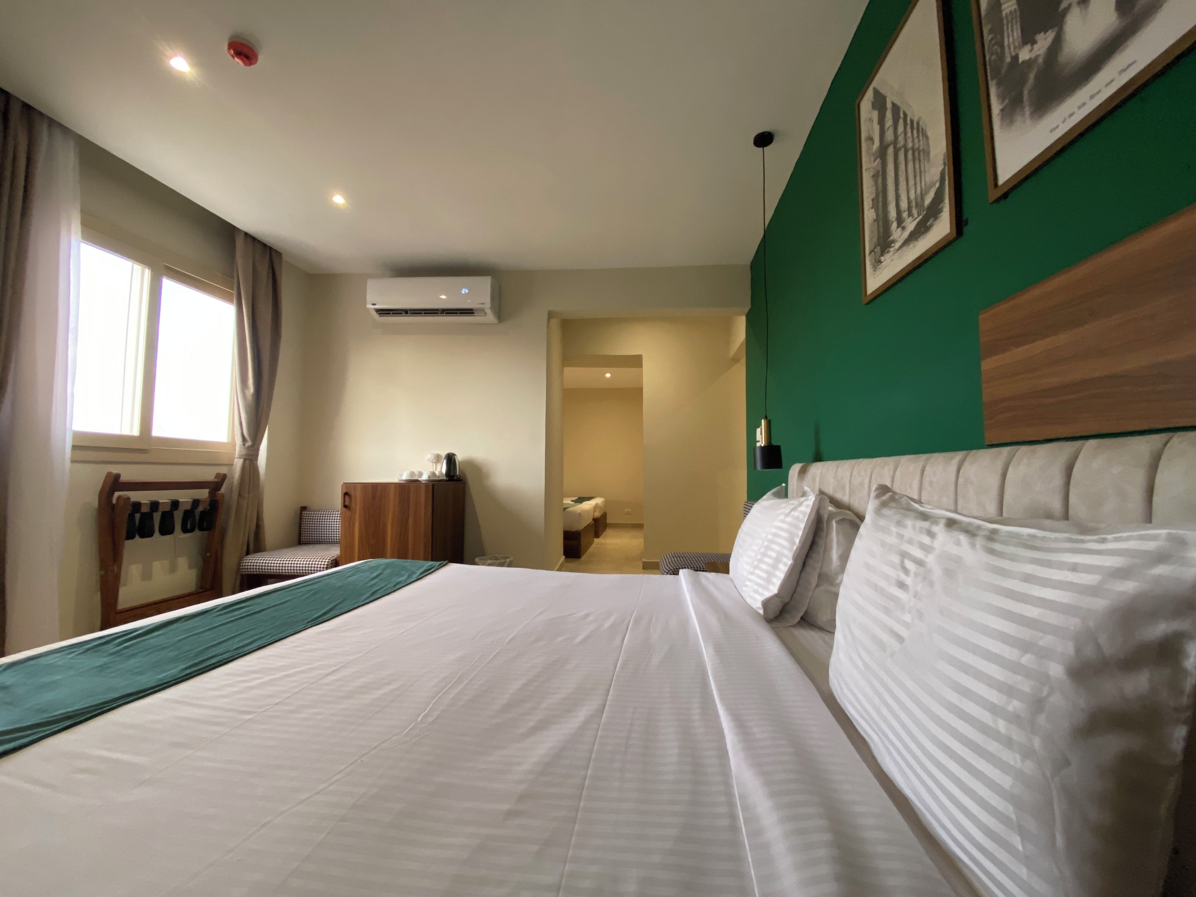 A hotel room with a large bed, a wooden headboard, a desk with a chair, a window with curtains, and a green accent wall.