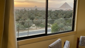 A room with a large window overlooking palm trees and the pyramids. - The Nomad Pyramids Boutique Hotel (Giza)