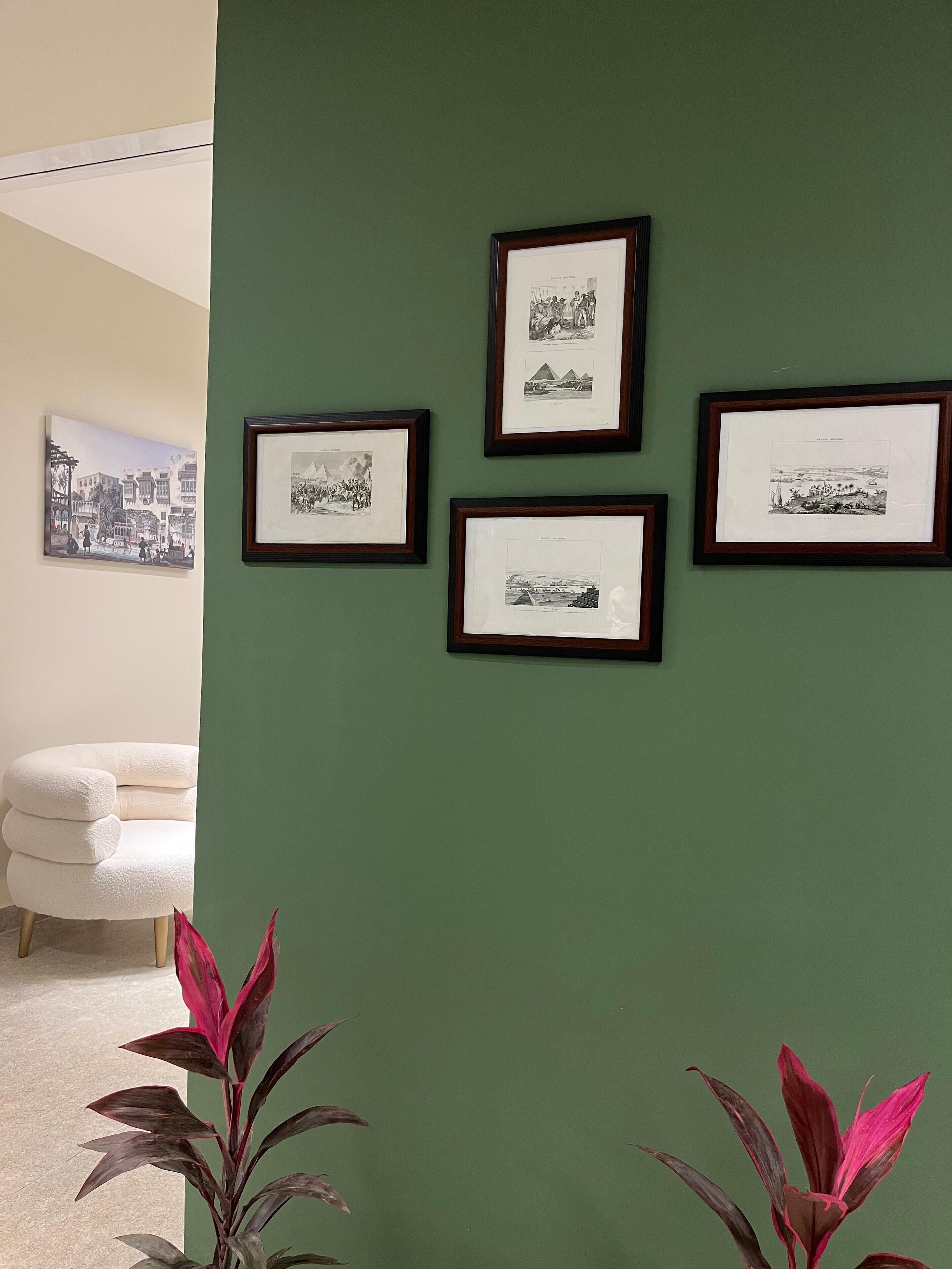 A room with a green wall, four framed black and white photographs, a white chair, and two potted plants with red leaves.