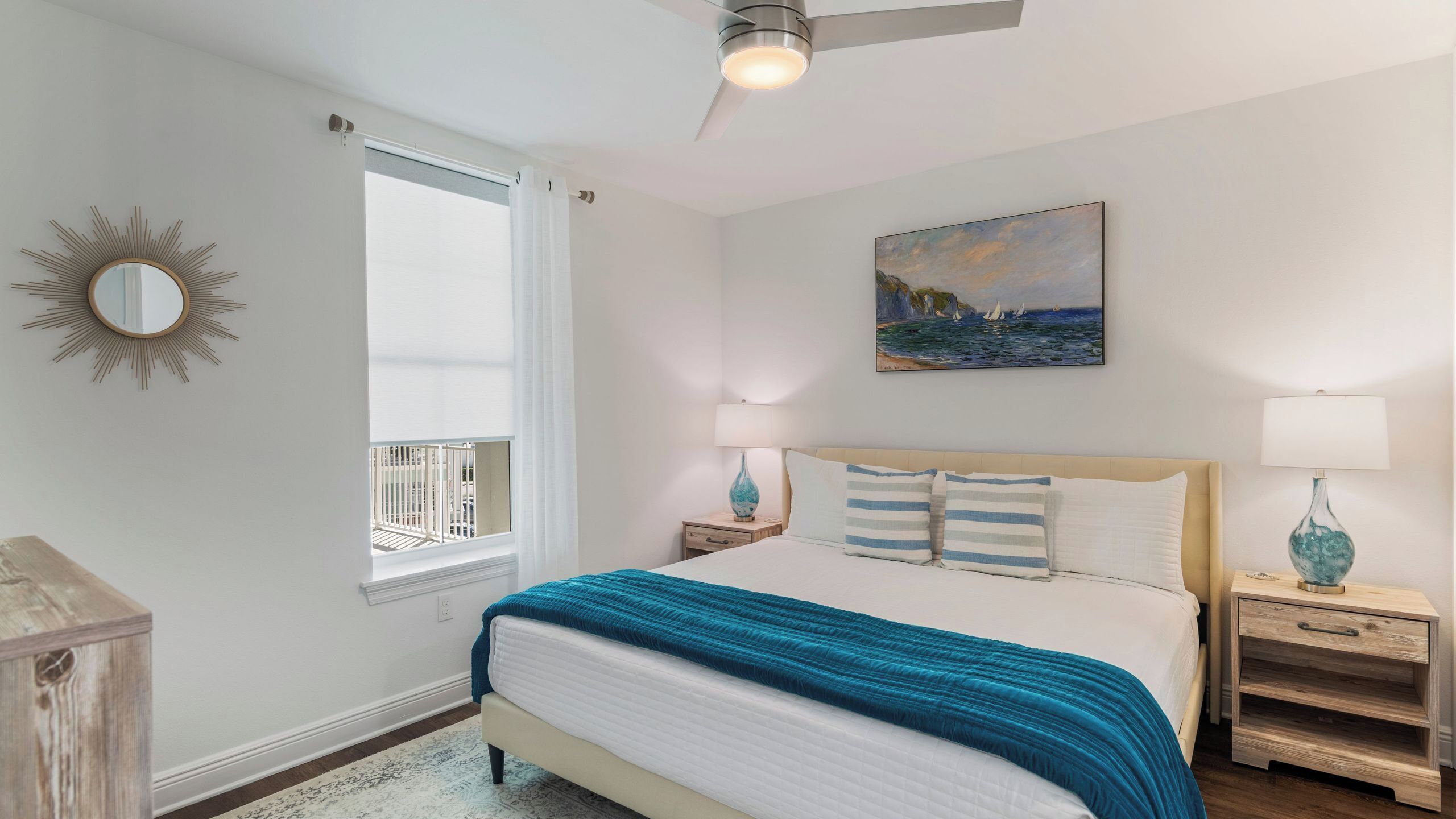 Quarto com cama, ventilador de teto, janela com persianas, quadro e abajures.