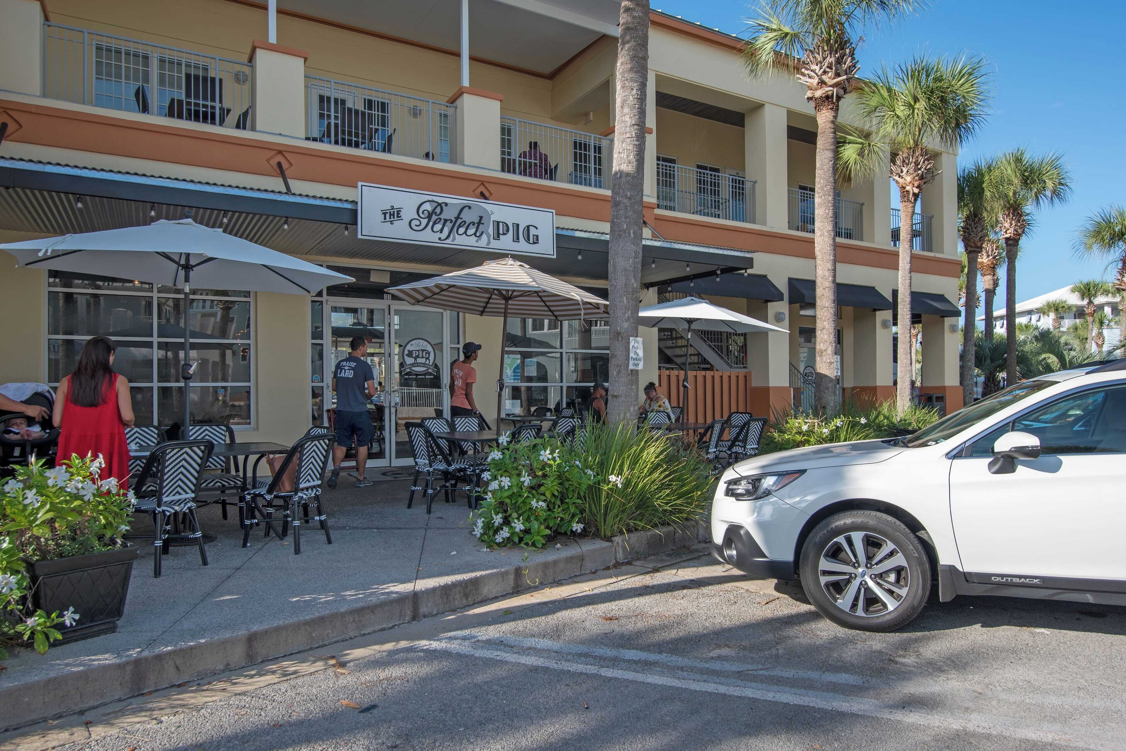 An outdoor seating area with tables and chairs, a white car parked nearby, and a building with a sign reading "The Perfect Pig".