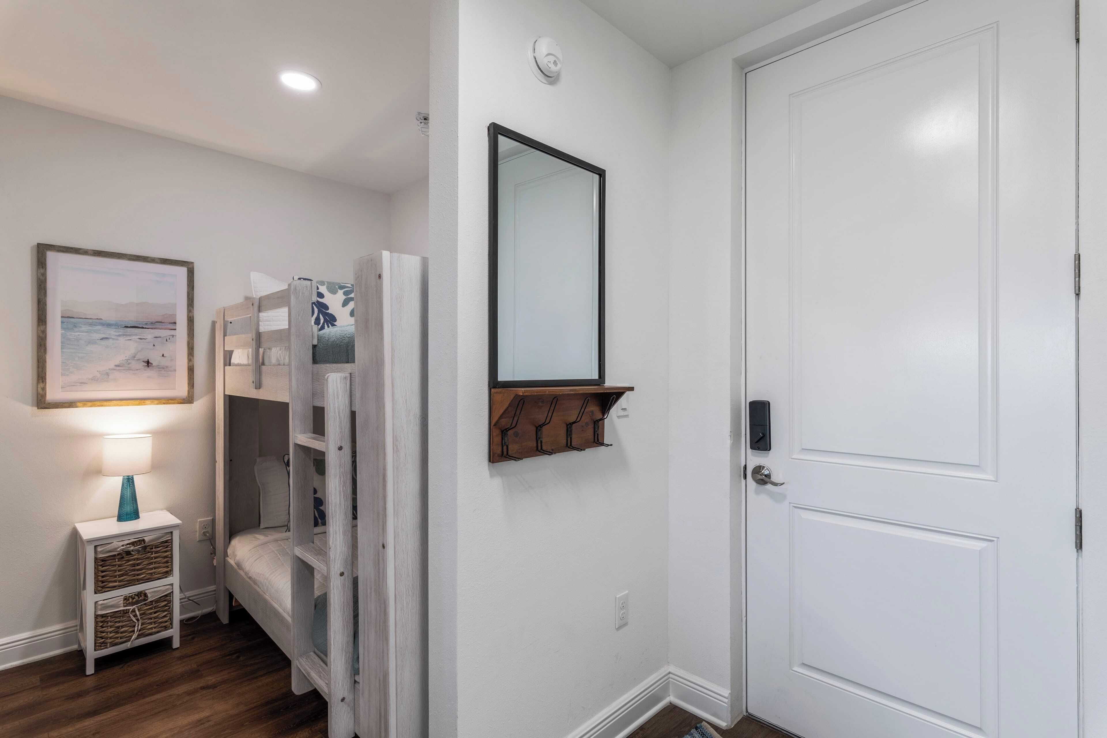 Une chambre avec un lit superposé, une table de chevet avec une lampe, un tableau encadré au mur, un miroir et une porte.