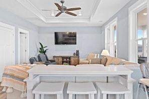 A modern living room with a sectional sofa, a flat-screen TV, a ceiling fan, and a dining area with white tables. - A Sunny Daze by Brunswick Vacation Rentals: Ocean Views, Elevator, Heated Pool (Ocean Isle Beach)