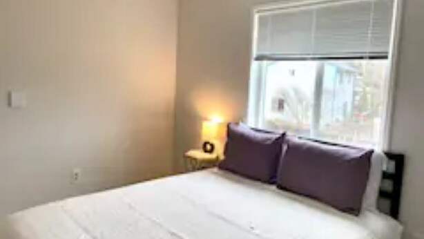 Un dormitorio con cama, mesita de noche, lámpara y ventana con persianas.