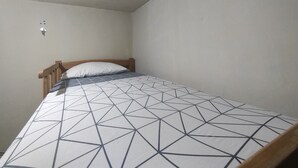 Ein kleines Zimmer mit einem Einzelbett, einer geometrisch gemusterten Bettdecke, einem hölzernen Kopfteil und einer schlichten weißen Wand.